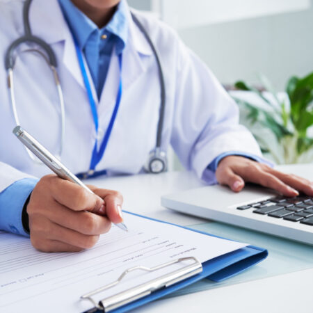 hands-unrecognizable-female-doctor-writing-form-typing-laptop-keyboard